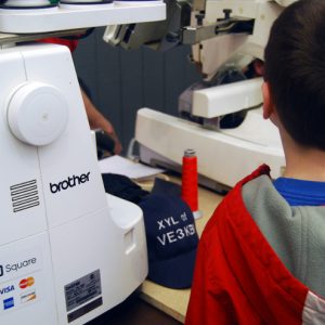 A young boy watches as Durham Hamfest vendor Sew Special Embroidery creates custom personalized hats and shirts for hams with their call signs.